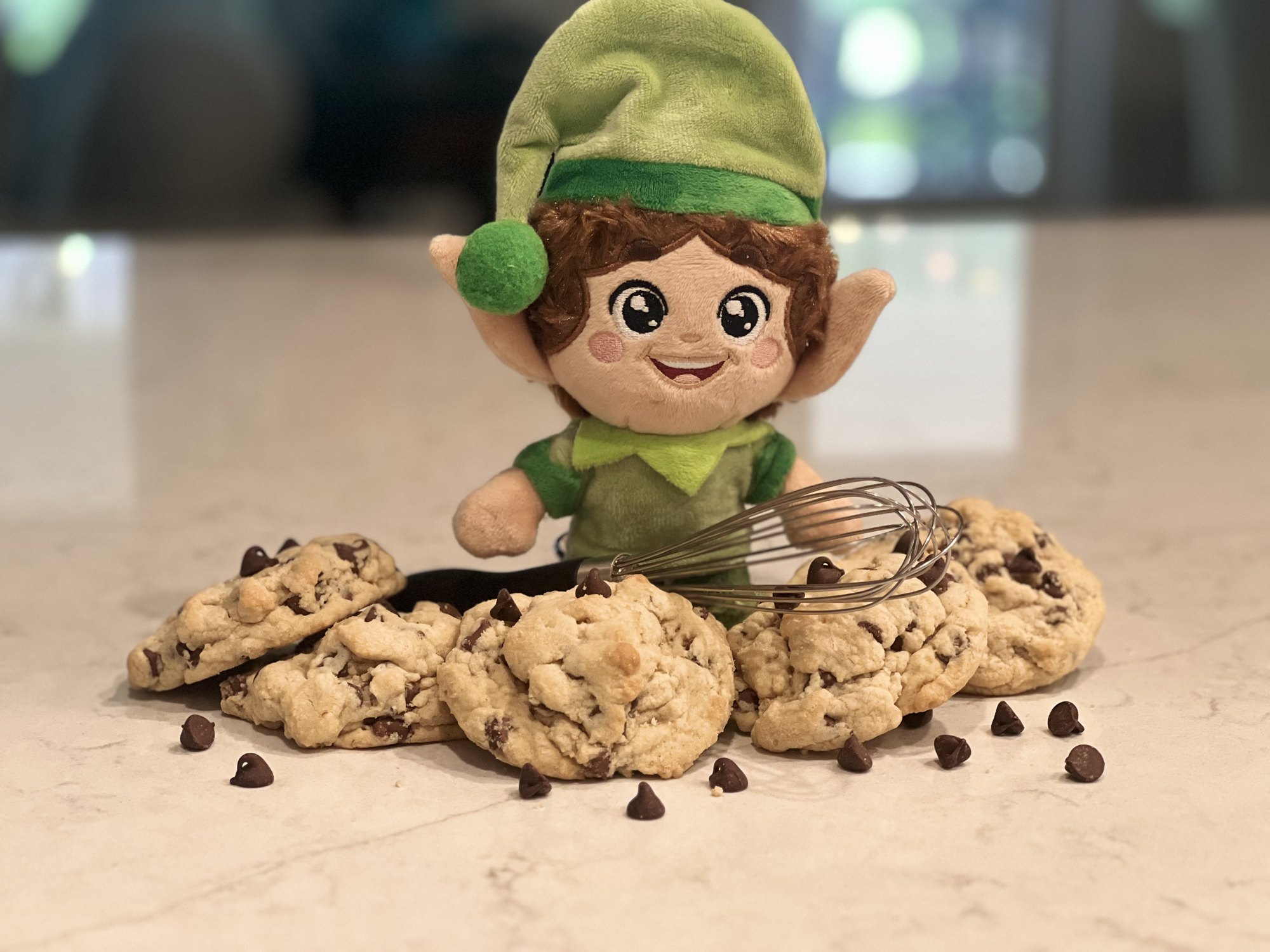 Green Kringle Elf behind a pile of chocolate chip cookies and a whisk