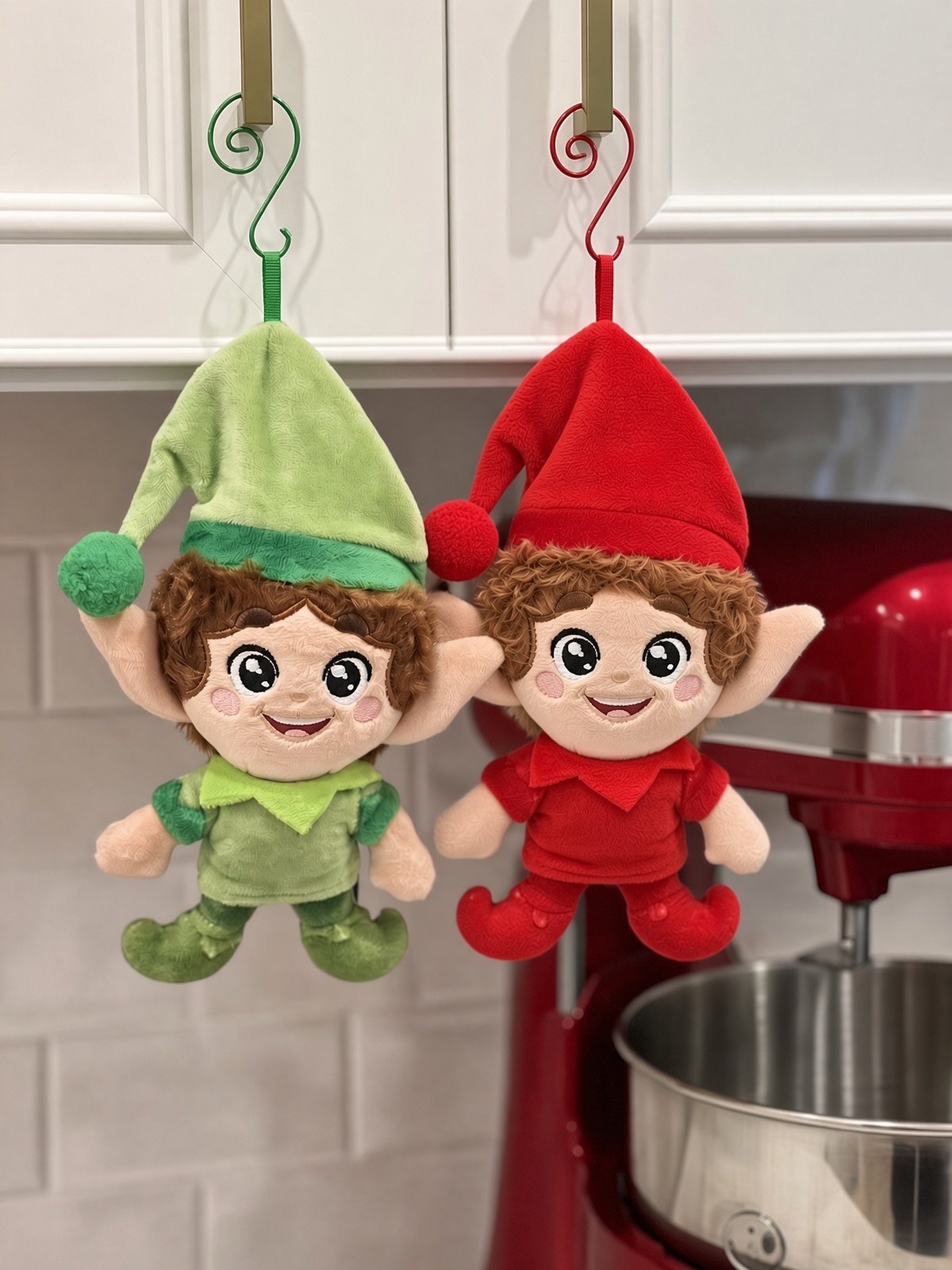 Green and red Kringle Elves hanging on kitchen cabinet hooks beside a red stand mixer